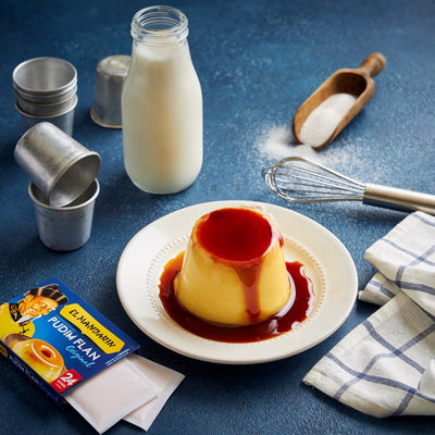 Creme caramel on a plate with milk bottle, measuring spoon, and whipped cream canisters on a blue surface.