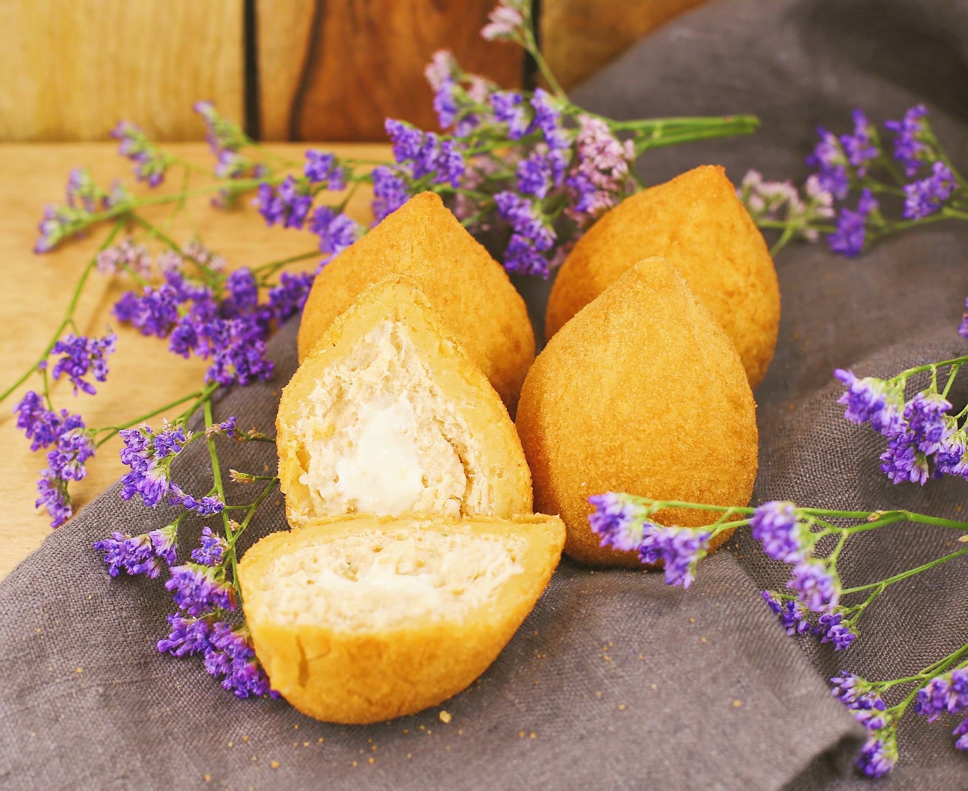 Three fried coxinhas with one cut open, surrounded by purple flowers on a dark surface.